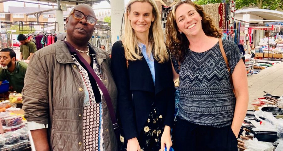 Aggie, a participant in the Fruit & Veg on Prescription project, stands smiling at Chrisp Street Market with BBC London journalist Victoria Cook and Alexandra Rose Charity’s Hannah Dakin. Stalls with fruit, vegetables, and textiles are visible in the background.