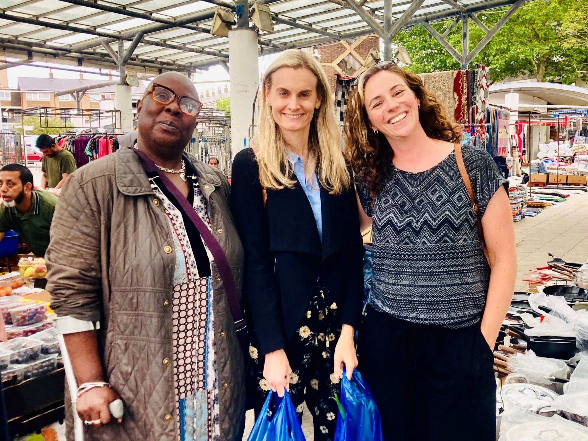 Aggie, a participant in the Fruit & Veg on Prescription project, stands smiling at Chrisp Street Market with BBC London journalist Victoria Cook and Alexandra Rose Charity’s Hannah Dakin. Stalls with fruit, vegetables, and textiles are visible in the background.