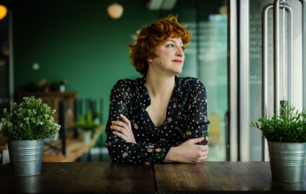 Corin sits at a wooden desk, with a green background. She is looking out of the window and smiling.