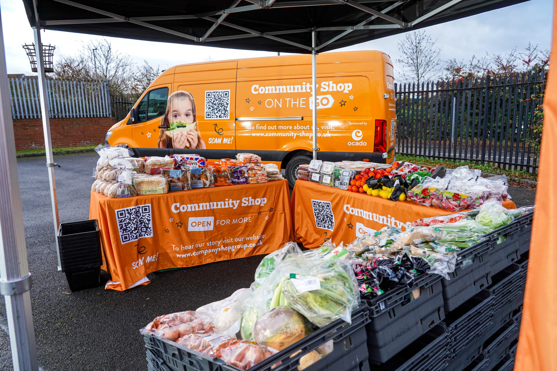Community Shop On the Go van and market stall offering fresh fruit, vegetables, bread, and groceries in a local neighbourhood.
