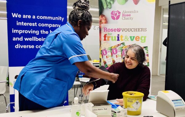 Nurse takes blood pressure of patient as part of the Fruit & Veg on Prescription launch in Newham. They both sit in front an Alexandra Rose banner.